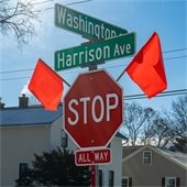 A red STOP sign with an “ALL WAY” plaque stands at the intersection of Washington Street and Harrison Avenue, with red safety flags attached to the signpost. Residential houses and leafless trees are visible in the background.